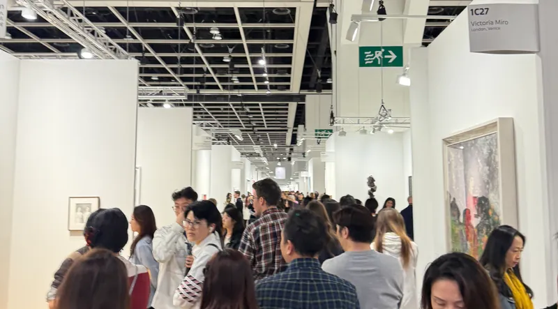 Art Basel Hong Kong 2025: Crowded art fair with people walking through a hallway of white booths. Art pieces are visible on walls.